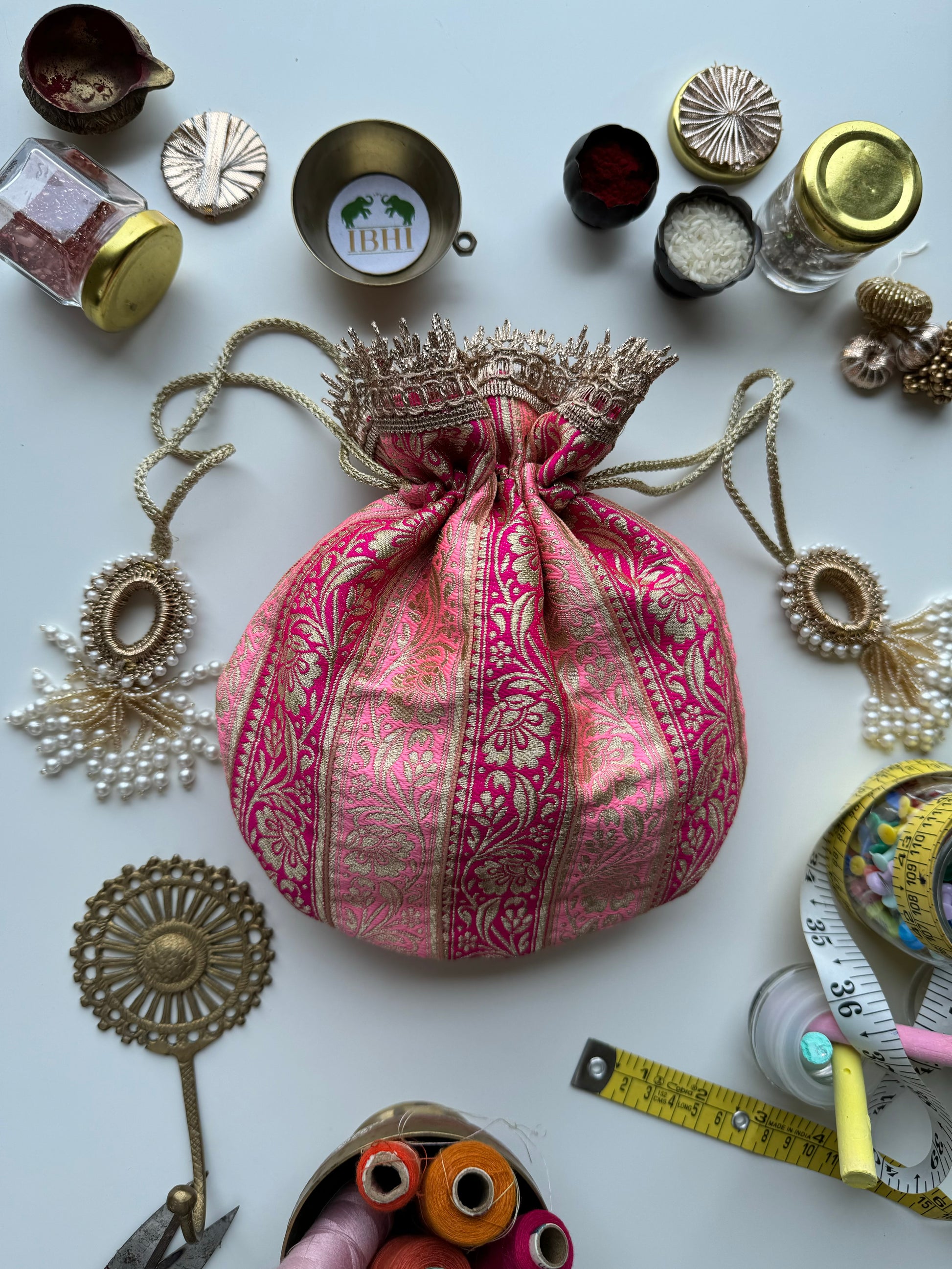 Royal pink Banarasi potli bag with gold zari crown lace and drawstring, surrounded by traditional festive decor items.